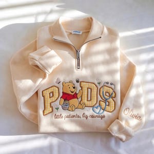 Peut inclure: Un sweat-shirt crème à fermeture éclair quart, orné d'une broderie de Winnie l'ourson, des lettres PDS en vichy jaune, un stéthoscope et le texte "little patients, big courage". Le prénom "Olivia" est brodé sur la manche.
