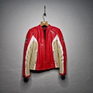 May include: A red, white, and beige leather motorcycle jacket featuring a mandarin collar with snap closures, zippered pockets, and the word "Texport" printed in white on the chest. The jacket has long sleeves with black zipper details at the cuffs.