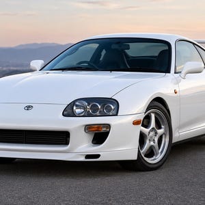 May include: A white Toyota Supra sports car featuring a sleek aerodynamic body, a prominent rear spoiler, silver five-spoke alloy wheels, and clear headlights with dark inner housings. The vehicle displays the Toyota emblem on the front hood.