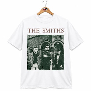 Może przedstawiać: Biały t-shirt z krótkim rękawem z czarno-białym nadrukiem graficznym przedstawiającym zespół The Smiths przed Salford Lads Club. Napis THE SMITHS jest wydrukowany brązową czcionką szeryfową nad członkami zespołu na grafice.