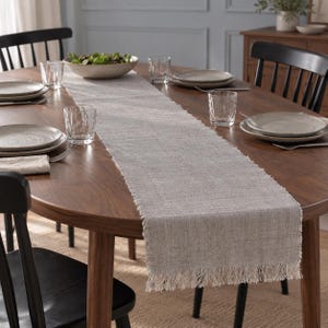 May include: A rectangular light gray table runner made of a woven fabric with a visible textured weave and frayed fringe edges. The runner is centered on a wooden dining table, providing a neutral base for place settings and decor.