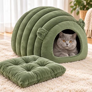 May include: A sage green velvet pet bed with a rounded, ribbed dome structure and two small ear-like accents on the top. A matching square, tufted velvet cushion sits in front of the bed, while a gray cat rests inside the arched opening.