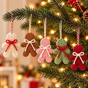 May include: Five crocheted gingerbread-style ornaments hanging from a pine tree. The figures are cream, brown, pink, green, and red, each featuring a small tied bow at the neck and a striped string loop for hanging on a branch.