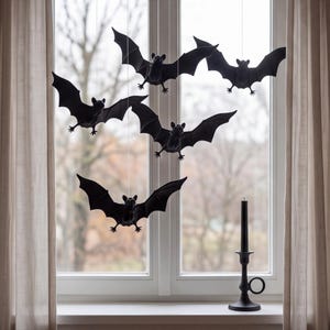 May include: Four black bat ornaments suspended by thin strings in front of a window, each featuring outstretched wings and pointed ears. A single black taper candle stands in a black candle holder on the windowsill below the hanging bats.
