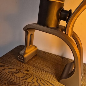 May include: A matte grey 3D-printed stand with a curved arch design supporting a cylindrical black device. The stand features a wide base with visible layer lines and a smooth finish, while the black device has a thin orange accent ring.
