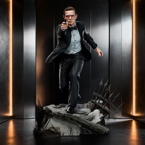 Pode incluir: Uma estatueta detalhada de uma pessoa com smoking preto, camisa branca e gravata borboleta, segurando uma pequena arma preta. A figura está sobre uma base rochosa texturizada com uma seção de cerca de metal quebrada e dinâmica.