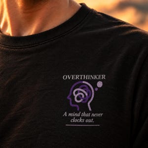 Pode incluir: Uma t-shirt preta de gola redonda com um gráfico no peito. O design inclui a palavra OVERTHINKER acima de um contorno roxo de uma cabeça humana com um nó emaranhado. Abaixo da cabeça está o texto A mind that never clocks out.