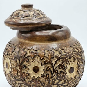 Puede incluir: Un tarro redondo de madera con tapa a juego, que presenta intrincados patrones florales tallados a mano. El diseño muestra tallos y hojas tallados en color marrón oscuro que contrastan con un fondo de madera natural clara y texturizada.