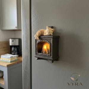 May include: A dark grey, rectangular wall-mounted decorative light shaped like a small stove with a glowing orange interior. A light tan cat figurine is sleeping on the top surface of the stove. The unit features a curved frame around the opening.