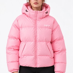 Könnte beinhalten: Eine leuchtend rosa Steppjacke mit hohem Kragen, einer Kapuze mit verstellbaren schwarzen Kordelzügen und einem durchgehenden Reißverschluss. Das Wort DRYP ist in Weiß auf der linken Brust aufgedruckt. Die Jacke hat lange Ärmel mit elastischen Bündchen.