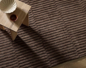 May include: A dark brown area rug with a repeating horizontal ribbed texture. A light-colored wooden side table sits on the rug, holding a white scalloped bowl and a small, round, dark brown ceramic dish on its flat surface.