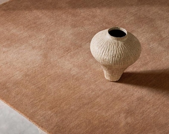 May include: A solid, light brown area rug with a short, dense pile texture. Resting on the rug is a cream-colored ceramic vase featuring a wide, rounded body, a narrow neck, and a deeply grooved, vertical ribbed texture across its entire surface.