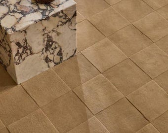 May include: A tan area rug featuring a grid of square tufted sections, positioned next to a cubic side table made of white and brown veined marble, which holds a small, round, dark brown polished stone bowl on its top surface.
