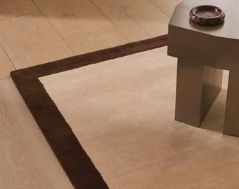 May include: A rectangular area rug featuring a large beige center panel framed by a wide, dark brown border. A solid, light brown geometric side table stands on the rug, topped with a small, round, dark brown marble-patterned dish.
