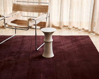 May include: A deep burgundy rectangular area rug with a paneled texture, featuring a chrome-framed lounge chair with beige fabric upholstery and a small, hourglass-shaped off-white side table placed on top of the rug surface.
