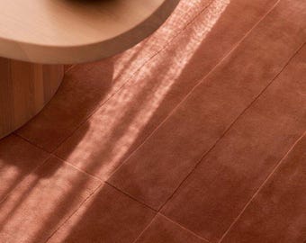 May include: A rectangular area rug in a solid terracotta color features a grid of stitched seams creating a panel effect. The rug is positioned beneath a light-toned wooden circular table holding a ceramic bowl with three orange persimmons.