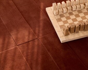 May include: A stone chess set with light beige and tan checkered squares and carved pieces, positioned on a deep rust-colored rug featuring a series of vertical seams and a soft, textured pile surface with subtle light reflections.