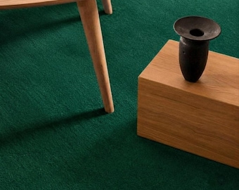 May include: A dark brown, wide-mouthed ceramic vase sits atop a rectangular light oak wooden block. Nearby, a portion of a light oak wooden table with a single visible leg is positioned, all set against a deep green carpeted floor.