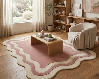 May include: A rectangular area rug featuring a wavy, scalloped edge design. The rug has a solid dusty rose center surrounded by concentric, undulating bands of cream and light pink, creating a soft, layered appearance on the wooden floor.