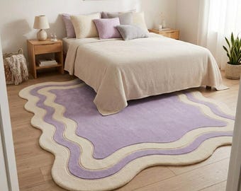 May include: An area rug with an irregular, wavy-edged shape featuring a large central lavender section surrounded by concentric cream and lavender wavy borders. The rug is placed on a light wood floor in front of a bed with a cream-colored quilt.