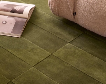 May include: A rectangular olive green area rug with a grid pattern of stitched seams, featuring a textured beige bouclé sofa cushion and an open book with black and white photography resting on the surface of the rug.