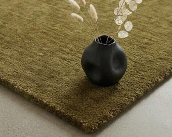 May include: A dark olive green textured area rug featuring a matte black ceramic vase with an irregular, rounded shape. The vase contains a collection of dried white bunny tail grass and translucent silver dollar plant stems.