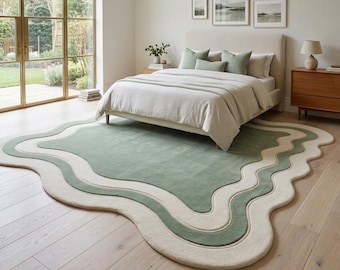 May include: An area rug with an irregular, wavy organic shape featuring a large sage green center section surrounded by concentric bands of cream and sage green. The rug has a soft, plush texture and is positioned on a light wood floor.