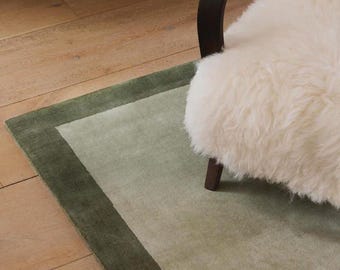 May include: A rectangular area rug featuring a light sage green center panel framed by a darker forest green border. The rug is positioned beneath a chair with a dark wooden armrest and a thick, white, long-haired sheepskin-style cover.