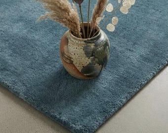 May include: A solid blue area rug with a thick, plush texture. Centered on the rug is a round, mottled ceramic vase featuring brown, cream, and dark gray tones, filled with dried pampas grass, lotus seed pods, and lunaria stems.