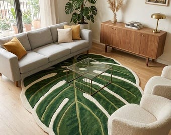May include: A large, green and white area rug shaped like a monstera leaf. The rug features a deep green center with lighter green veins and a thick, cream-colored border, positioned on a light wood floor beneath a glass-topped coffee table.