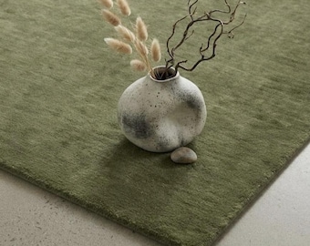 May include: A solid olive green area rug with a textured surface. Placed on the rug is a round, speckled off-white ceramic vase containing several beige dried bunny tail grasses and a dark brown twisted branch, with a small smooth stone.