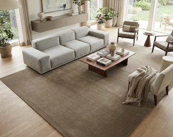 May include: A large rectangular area rug in a solid dark taupe color, featuring a smooth, low-pile texture. The rug is positioned centrally in a living room, covering a significant portion of the light-colored wooden floor beneath a sofa and chairs.
