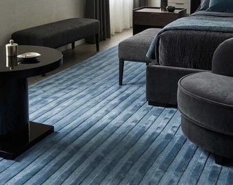 May include: A large area rug featuring a textured, linear pattern in varying shades of blue. The rug has a soft, plush pile with raised horizontal stripes that run across its surface, creating a consistent, repeating ribbed design throughout.