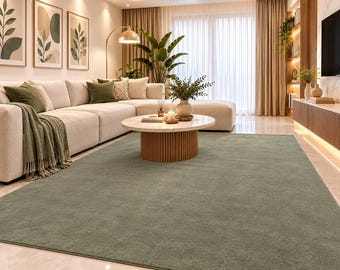 May include: A large, rectangular area rug in a solid sage green color with a soft, textured pile. The rug is positioned on a polished light-colored floor in front of a cream-colored sectional sofa, covering a significant portion of the floor space.
