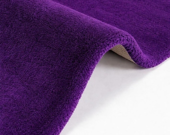 May include: A deep purple area rug with a soft, plush, and textured surface. The rug is folded over to reveal a light beige backing material. The fabric has a dense, uniform pile that creates a consistent color appearance across the entire surface.
