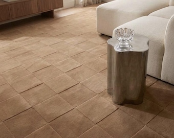 May include: A large, square-patterned tan area rug covering the floor, featuring a textured, grid-like design. The rug is positioned in a room with a light-colored sofa, a metallic side table, and a wooden media console in the background.