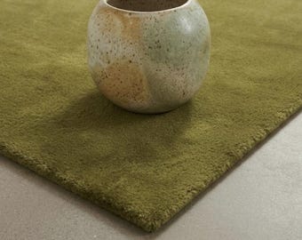 May include: A round, speckled ceramic vase with a mottled finish in shades of cream, sage green, and soft orange, resting on a solid olive green rug with a short, dense pile texture that covers the lower portion of the frame.