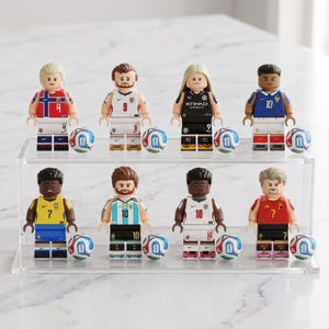 May include: Eight football player minifigures arranged on a two-tier clear acrylic display stand. Each figure wears a unique national or club team jersey with a corresponding number and is accompanied by a small, round, white and blue football.