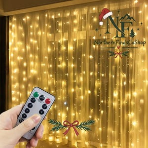 May include: A curtain of warm white LED string lights hanging in vertical strands, creating a glowing backdrop. A small white remote control with multiple buttons is held in the foreground to adjust the light settings and patterns.