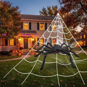May include: A large, black, fuzzy spider with glowing red eyes is positioned on a white, triangular spider web decoration. The spider has eight long, articulated legs and is displayed on a lawn in front of a house decorated for Halloween.