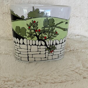 Puede incluir: Una maceta de cerámica blanca con una ilustración de paisaje pintada a mano. El diseño muestra un muro de piedra en primer plano, un árbol frutal con manzanas rojas, colinas verdes y una pequeña vaca blanca y negra a lo lejos.