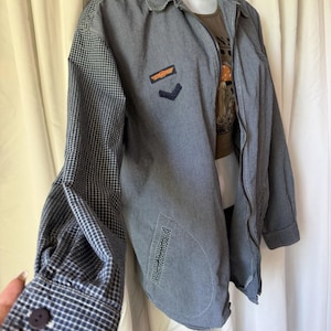 May include: A blue and white pinstriped zip-up jacket featuring a chequered pattern on the sleeves and shoulders. The chest displays two embroidered patches, one orange and one navy blue. The jacket includes a collar, long sleeves with buttoned cuffs, and two front pockets with chequered trim.