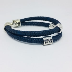 以下が含まれることがあります： 銀色のチャームビーズが付いた、2連の青い革のブレスレット。1つのビーズには、棒人間と家の家族のデザインが施されています。