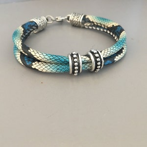 Puede incluir: Un brazalete de doble cadena con un patrón de piel de serpiente azul y blanco. El brazalete tiene detalles de metal plateado y un cierre.