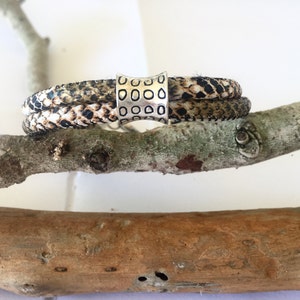 May include: A brown and white patterned leather bracelet with a silver metal bead. The bead has a textured design.