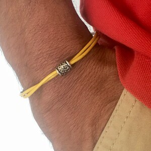 Könnte beinhalten: Ein gelbes Lederarmband mit einer silbernen Perle. Das Armband wird am Handgelenk getragen.