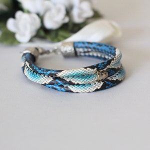 Op de afbeelding: Een drievoudig gewikkelde armband gemaakt van blauw en wit leder met een slangenhuidpatroon, een zilveren sluiting en een kleine gouden kraal.