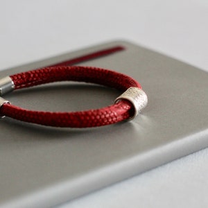 Könnte beinhalten: Ein rotes geflochtenes Armband mit einer silbernen Metallperle. Das Armband liegt auf einer grauen Oberfläche.