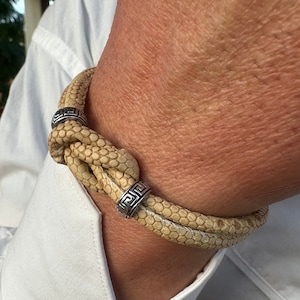 Puede incluir: Un brazalete de cuero marrón claro con un diseño de nudo y detalles metálicos plateados. El brazalete tiene un patrón geométrico en los detalles metálicos.