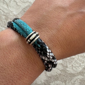 May include: A black and white snakeskin bracelet with a turquoise snakeskin accent and a silver metal clasp.